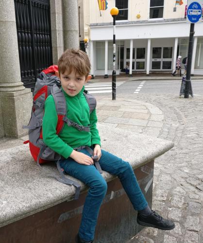Jowan with backpack, jeans and green Camber fleece top sat on a bench outside the Market House Penzance